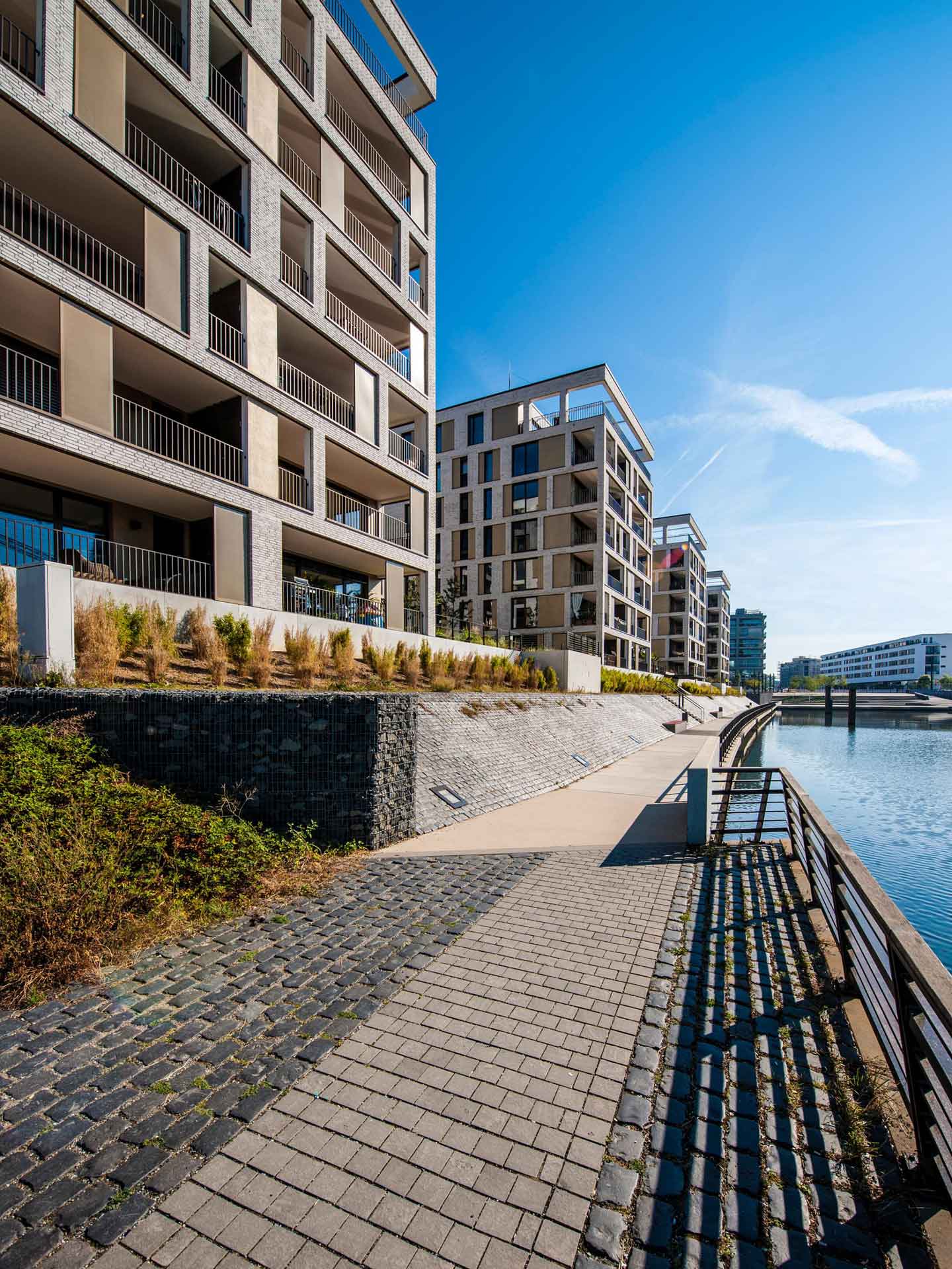 Eine moderne städtische Uferentwicklung mit Wohngebäuden. Die Architektur zeichnet sich durch klare Linien und große Fenster aus, mit Balkonen, die auf das Wasser blicken. Ein gepflasterter Weg verläuft entlang des Wassers, gesäumt von einer niedrigen Mauer und Landschaftsgestaltung mit Ziergräsern.