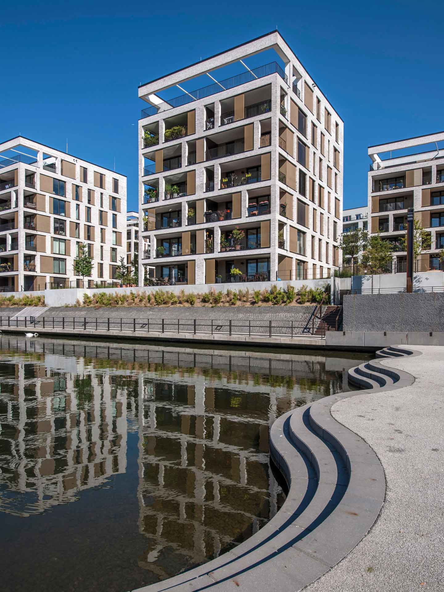 Moderne Wohngebäude mit mehreren Balkonen und großen Fenstern, die sich im Wasser eines Kanals oder Flusses im Vordergrund spiegeln. Die Architektur ist zeitgenössisch mit kubischem Design, und darüber spannt sich ein klarer blauer Himmel.