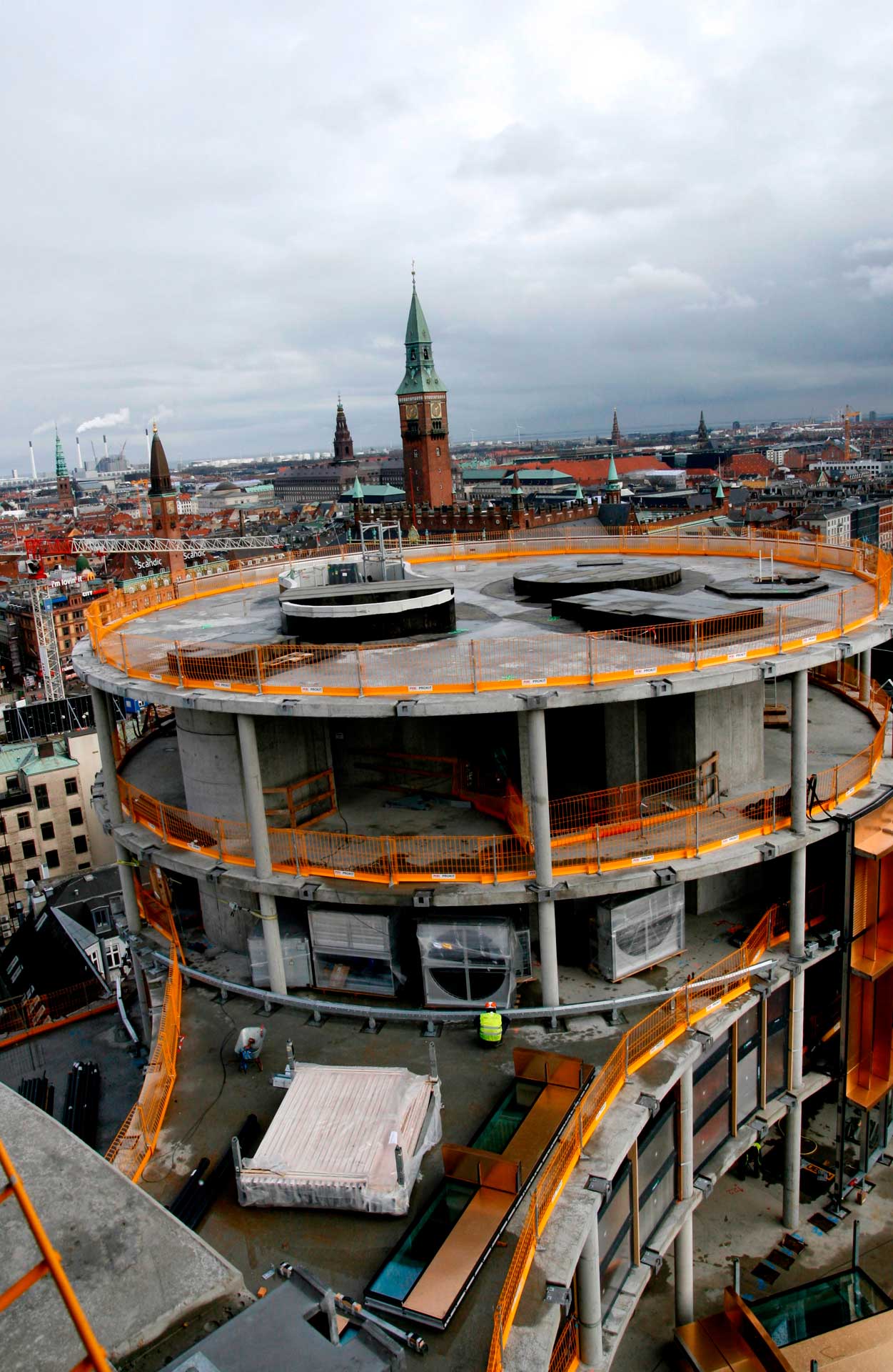 Eine große, mehrstöckige runde Baustelle mit orangefarbenen Sicherheitsbarrieren. Die Baustelle befindet sich in einer städtischen Umgebung mit Gebäuden im Hintergrund, darunter ein markanter Turm mit grünem Dach und Spitze.