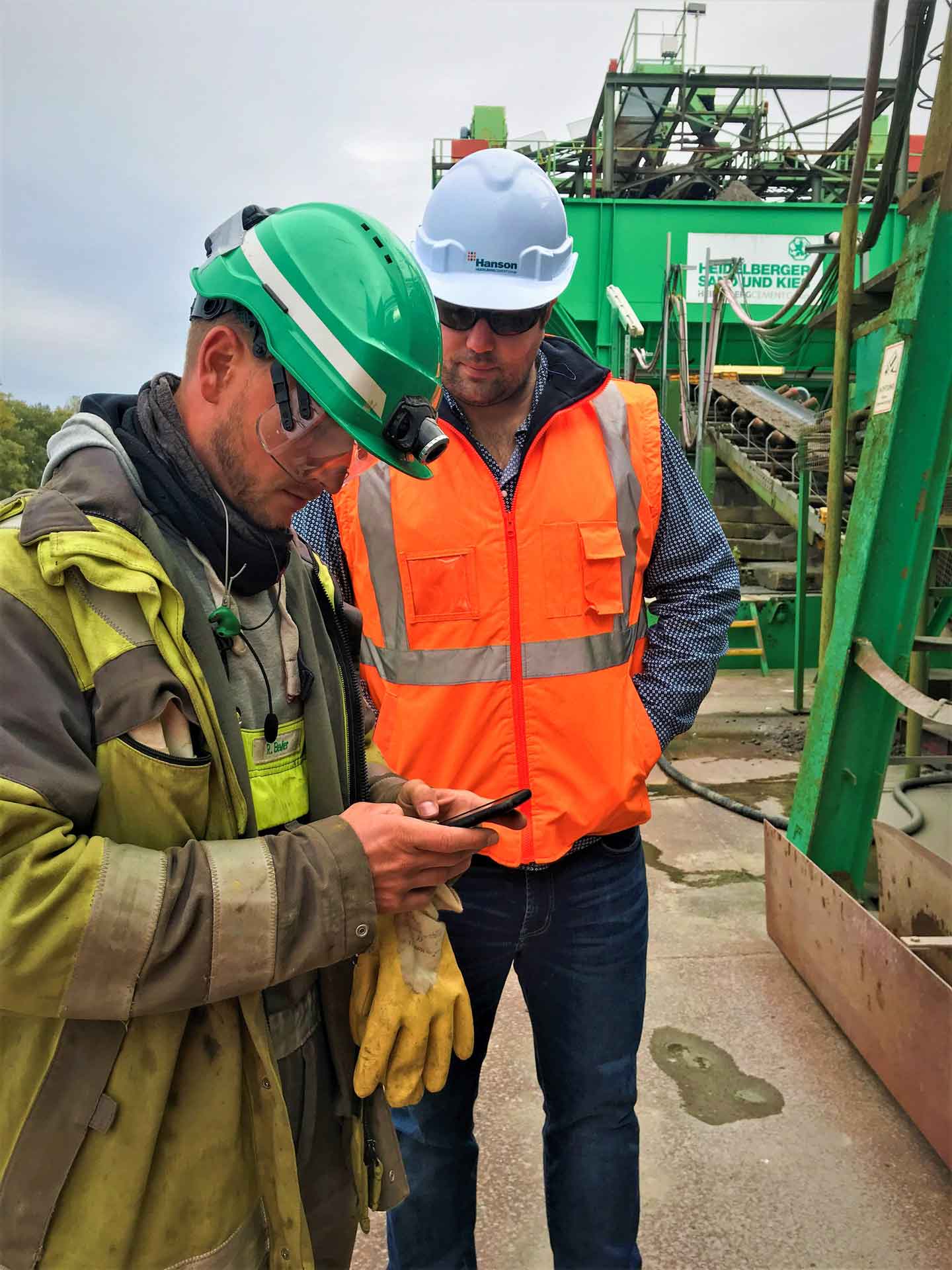 Two workers in safety gear discussing over a walkie-talkie at a construction site with heavy machinery in the background.