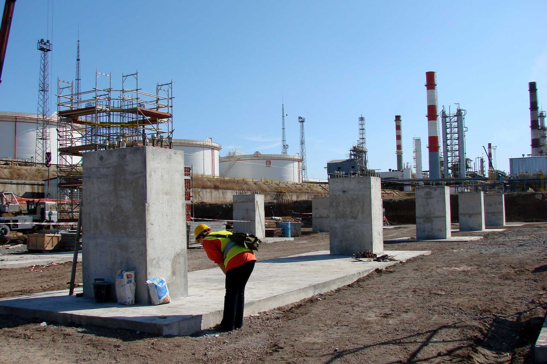 An industrial setting with multiple large cylindrical storage tanks and tall chimneys emitting no visible emissions against a clear sky. In the foreground, a person wearing high visibility clothing and a hard hat stands with their back to the camera, seemingly working or inspecting something on a small structure.