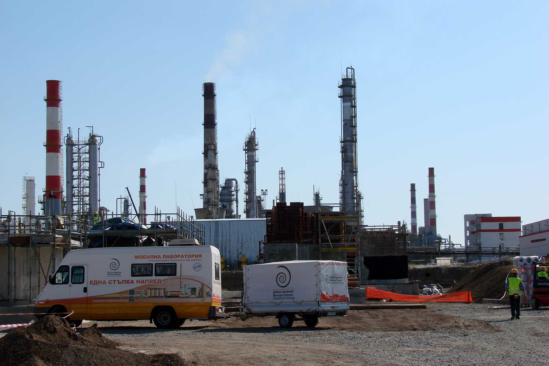 An industrial setting with multiple tall structures such as chimneys and distillation columns, indicative of a refinery or chemical plant. In the foreground, there are vehicles including a white van and a trailer, and two individuals wearing high-visibility vests, indicating construction or maintenance activity.