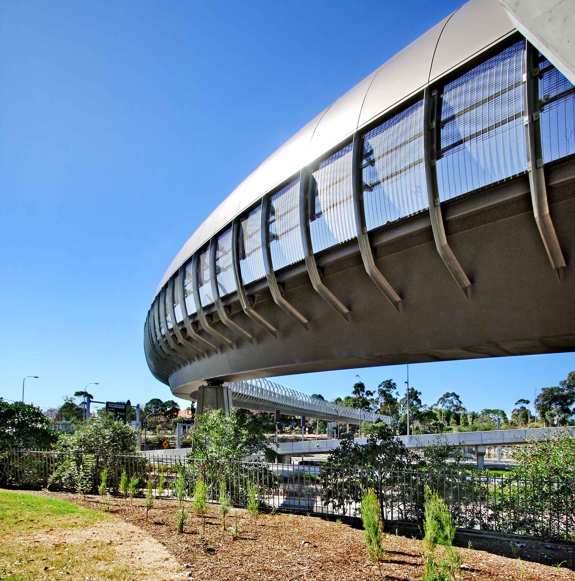 Eine moderne Fußgängerbrücke mit einem markanten, geschwungenen Design und einem überdachten Gehweg mit Metallgitterseiten. Die Brücke überspannt ein Gebiet mit Vegetation und befindet sich in einer städtischen Umgebung mit Gebäuden und klarem Himmel im Hintergrund.