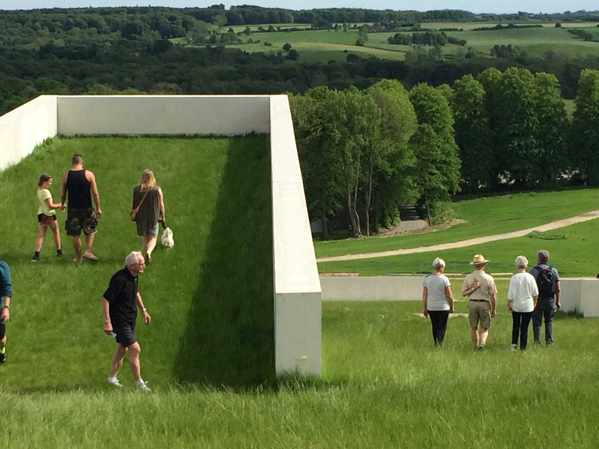 A group of people walk up and around a unique outdoor structure that looks like a grassy ramp. This structure is located in a green, open landscape with trees in the background.