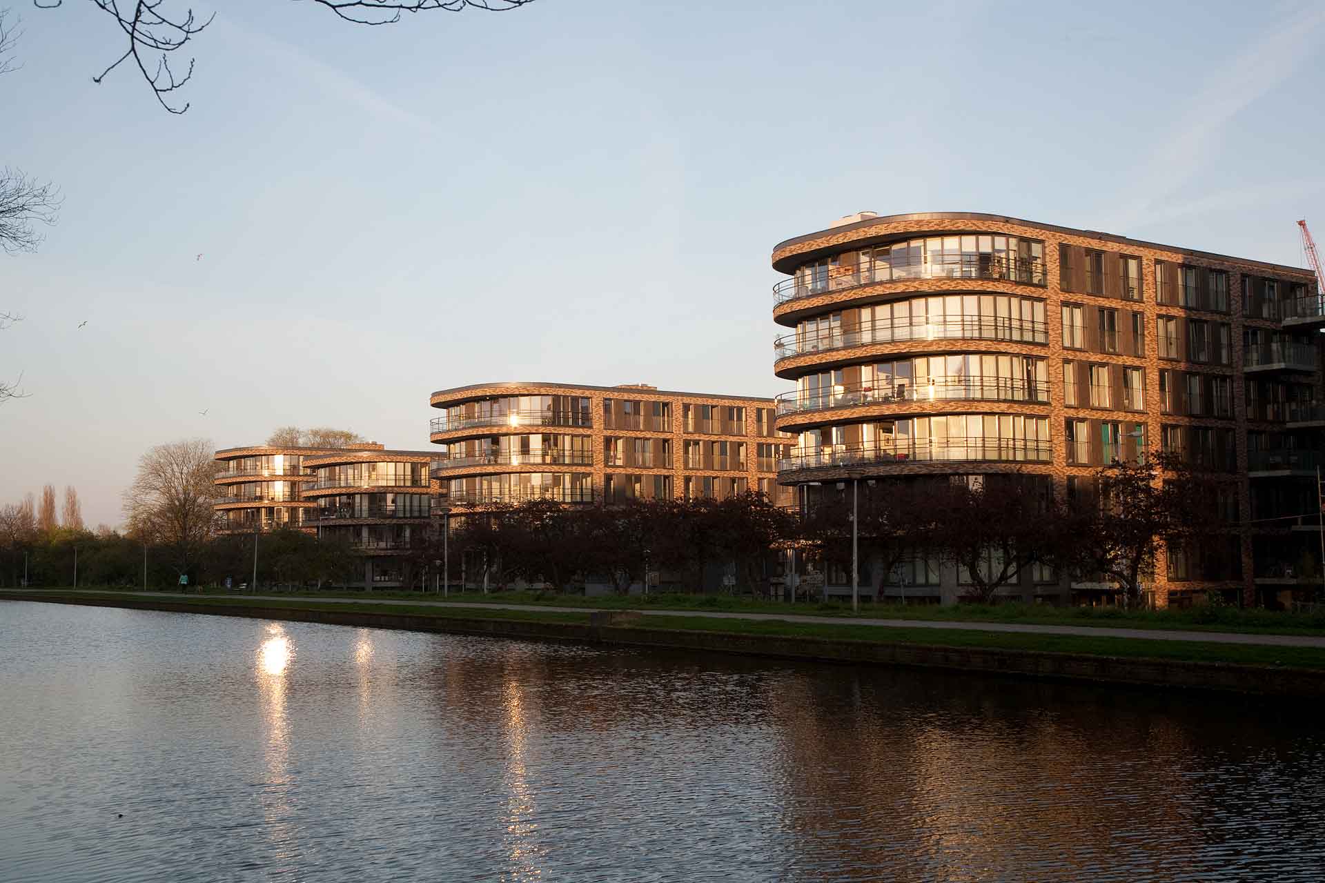 Eine Reihe moderner, mehrstöckiger Gebäude mit abgerundeten Fassaden und großen Glasfenstern, die das Sonnenlicht reflektieren. Diese Gebäude befinden sich neben einem ruhigen Gewässer, das die Strukturen und die umliegenden Bäume widerspiegelt.