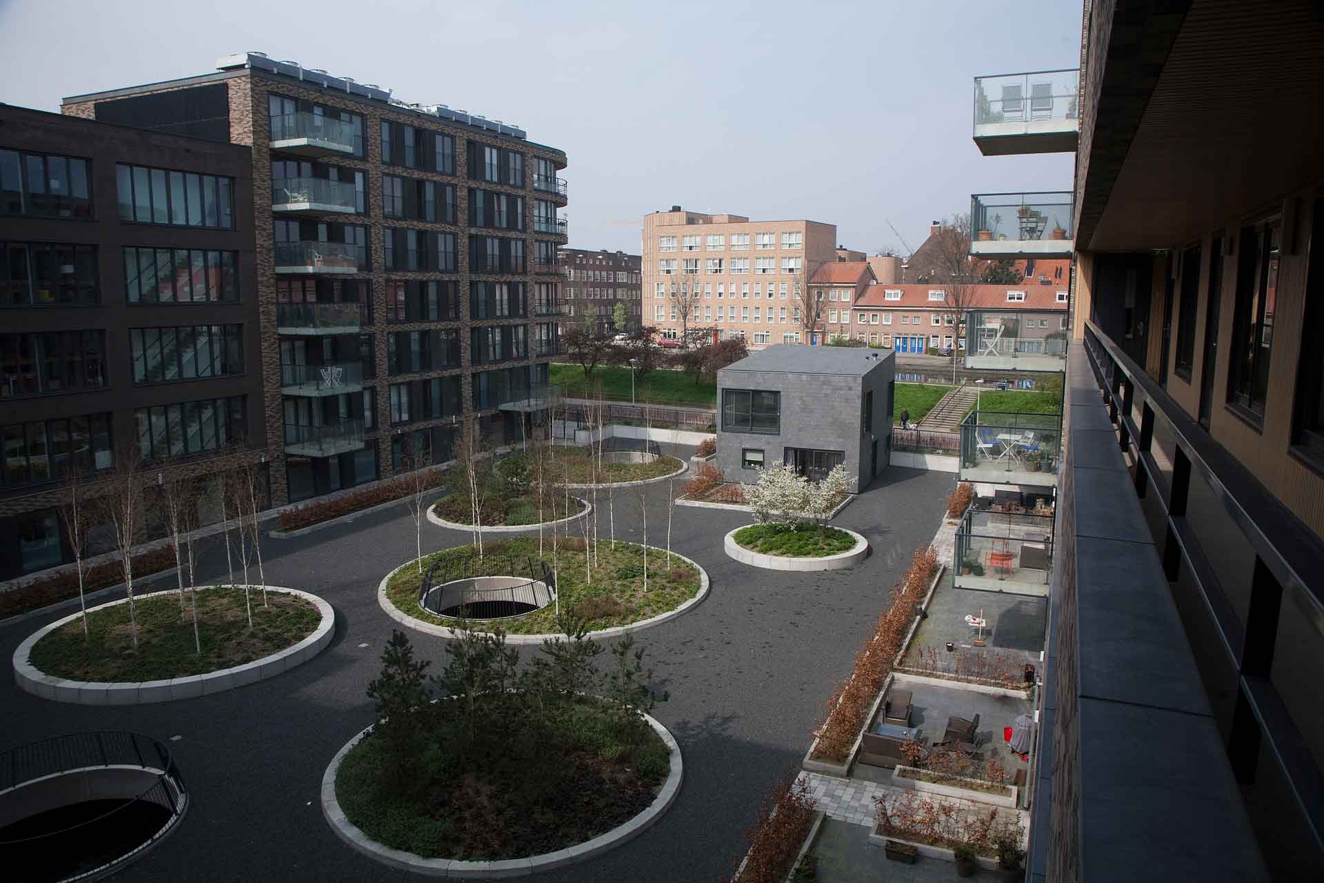 Ein urbanes Landschaftsbild, gesehen von einem Balkon. Es gibt moderne Gebäude mit großen Fenstern und einen Innenhofbereich mit kreisförmigen Grünflächen, Bäumen und Wegen.