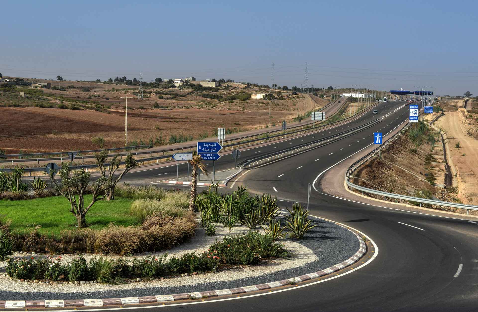 Eine mehrspurige Autobahn, die nach rechts abbiegt, unter klarem blauem Himmel mit trockenem Land auf beiden Seiten. Es gibt Straßenschilder, die Richtungen zu Städten oder Orten anzeigen, und im Vordergrund befindet sich ein Kreisverkehr mit Begrünung in der Mitte.