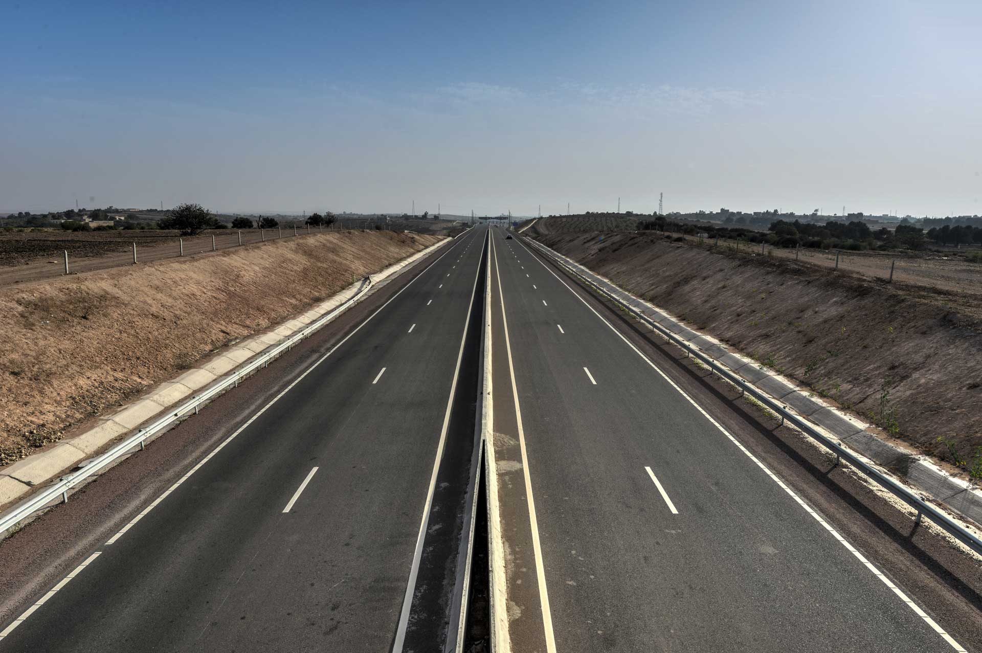 A straight, empty highway with multiple lanes extends into the horizon under a clear sky. The road is flanked by flat, barren land on both sides, with no vehicles or people visible.