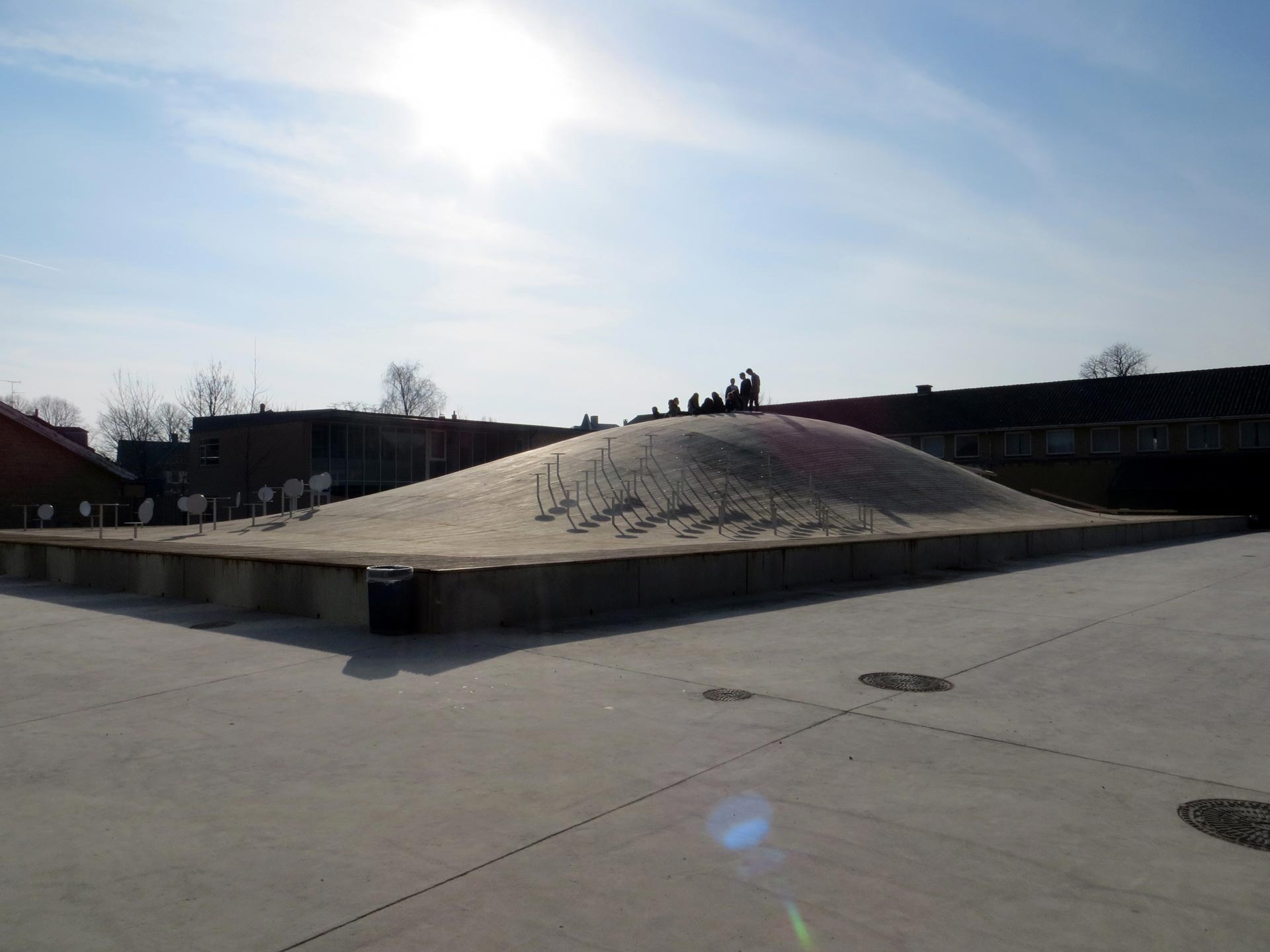 Ein Betondom mit glatter Oberfläche erhebt sich im Zentrum eines gepflasterten Platzes, umgeben von niedrigen Gebäuden unter einem klaren Himmel. Mehrere Personen stehen auf der Kuppel und werfen Schatten auf die gewölbte Fläche.