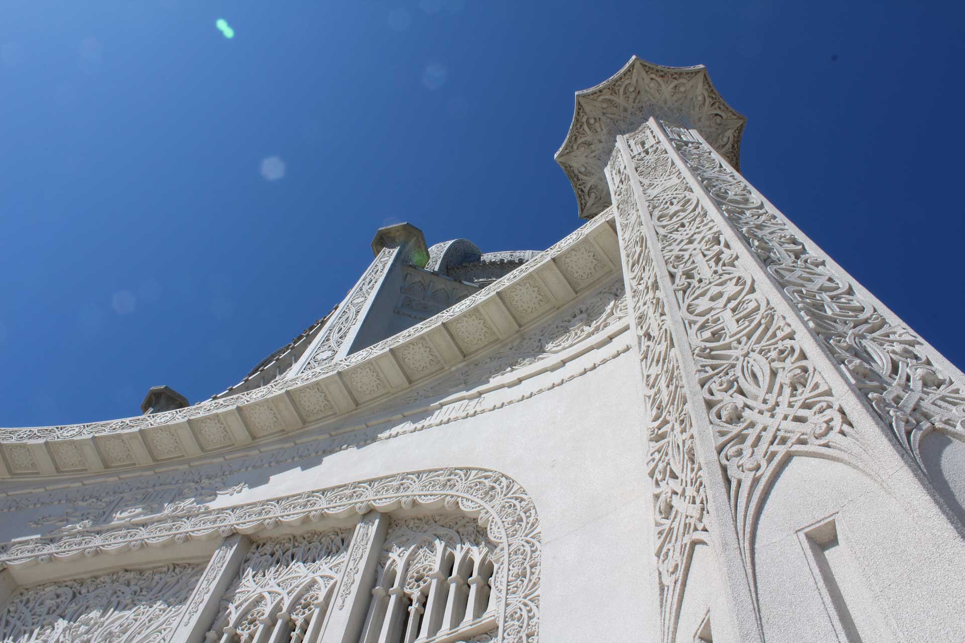 A close-up view of an intricately designed building, looking upwards towards the sky. The architecture features detailed patterns on its surface, with a clear blue sky in the background.