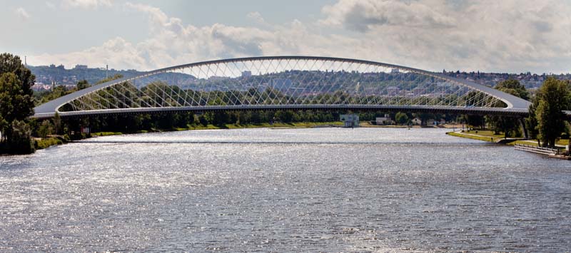 Modern bridge with a distinctive arch design spanning over a wide river. The arch has a lattice structure creating an intricate pattern against the sky. Sunlight reflects on the water below, with trees and buildings visible in the distance under a partly cloudy sky.