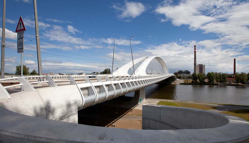 Modern bridge with a distinctive arch design, featuring a white framework and railings, spanning over a body of water under a clear blue sky with some clouds. An industrial chimney is visible in the distance on the right.