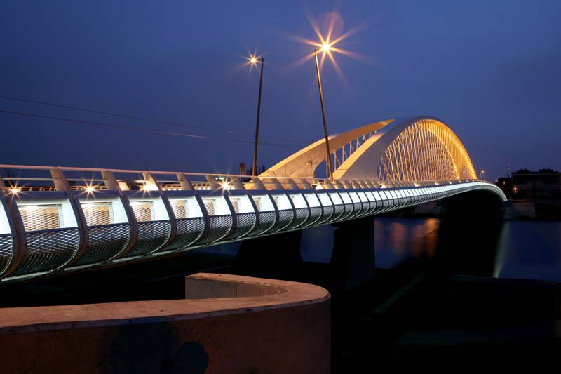 Modern bridge at twilight or night, illuminated by multiple lights. The bridge features a distinctive arching structure with repeating patterns. Starburst light effects are visible, with reflections on the water below.