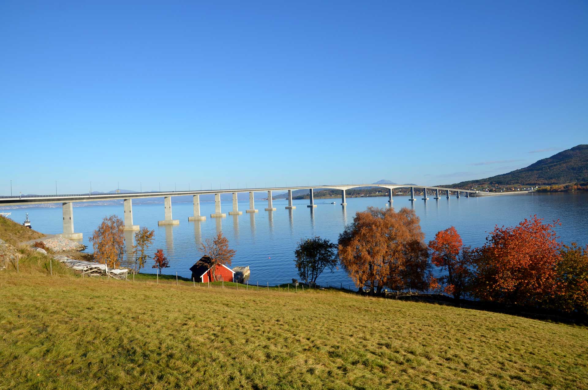 Eine lange Brücke überspannt ein großes Gewässer. Im Vordergrund befindet sich eine grasbewachsene Fläche mit Bäumen in Herbstfarben und einem kleinen roten Haus mit schwarzem Dach am Ufer.