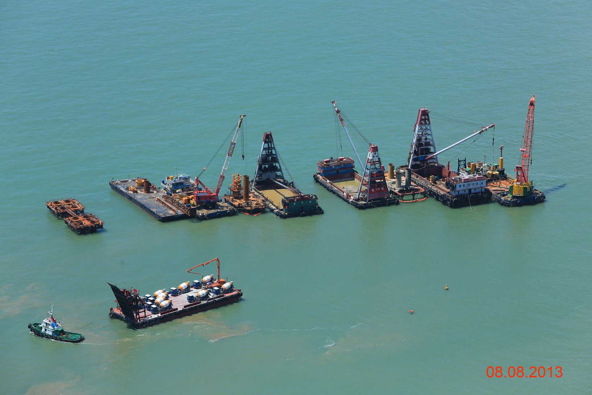 Maritime Baustelle mit mehreren Schiffen und Kränen auf dem Wasser, die auf ein Offshore-Bauprojekt hinweisen. Mehrere Lastkähne mit Kränen, einige stationär, andere von Schleppern manövriert, in hellgrün-blauen Küstengewässern. Datum ‘08.08.2013’ in der unteren rechten Ecke sichtbar.