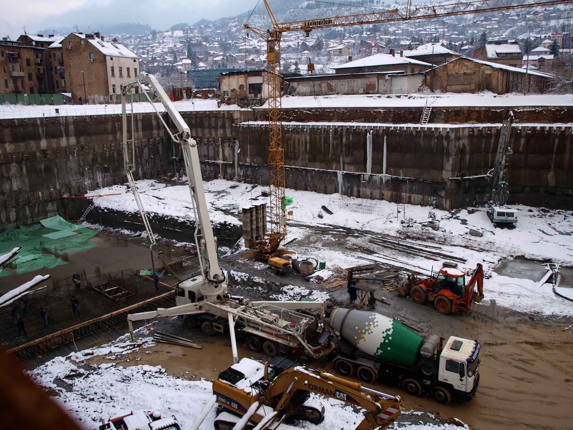 Eine Baustelle im Winter, erkennbar an der Schneedecke. Es gibt verschiedene Baufahrzeuge und -geräte, darunter ein Kran, ein Fahrmischer und ein Bagger.