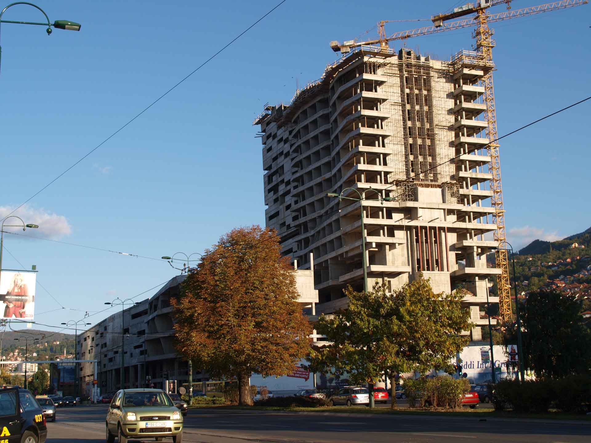 Eine Baustelle für ein großes Gebäude mit mehreren Stockwerken, sichtbaren Betonrahmen und einem Kran, der am Gebäude befestigt ist, was auf laufende Bauarbeiten hinweist. Im Vordergrund befinden sich kleinere Gebäude, Fahrzeuge auf der Straße und ein Hügel mit Häusern im Hintergrund.