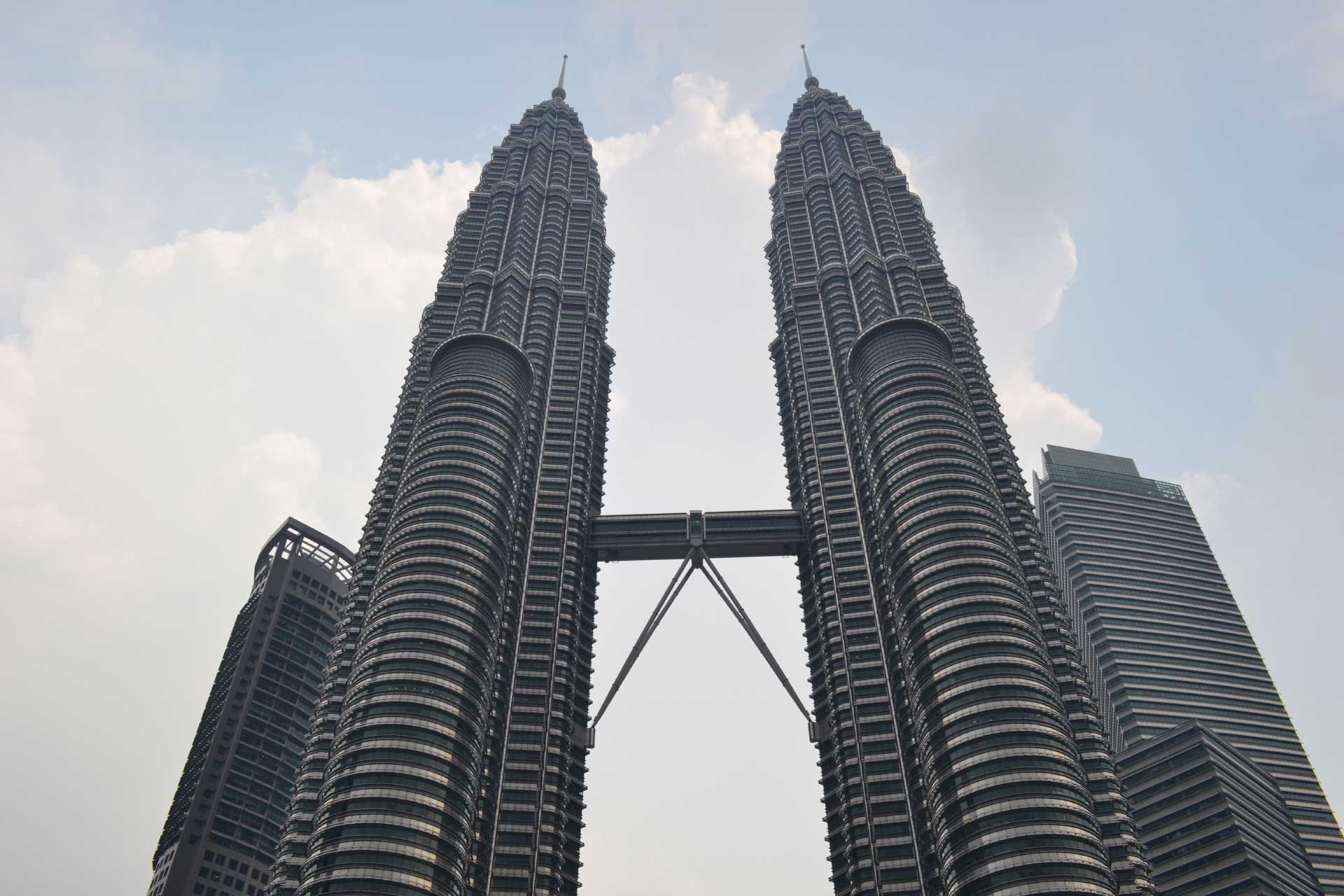 Two tall, symmetrical skyscrapers connected by a bridge span under a cloudy sky. The structure is made of concrete and glass and stands prominently against the skyline.