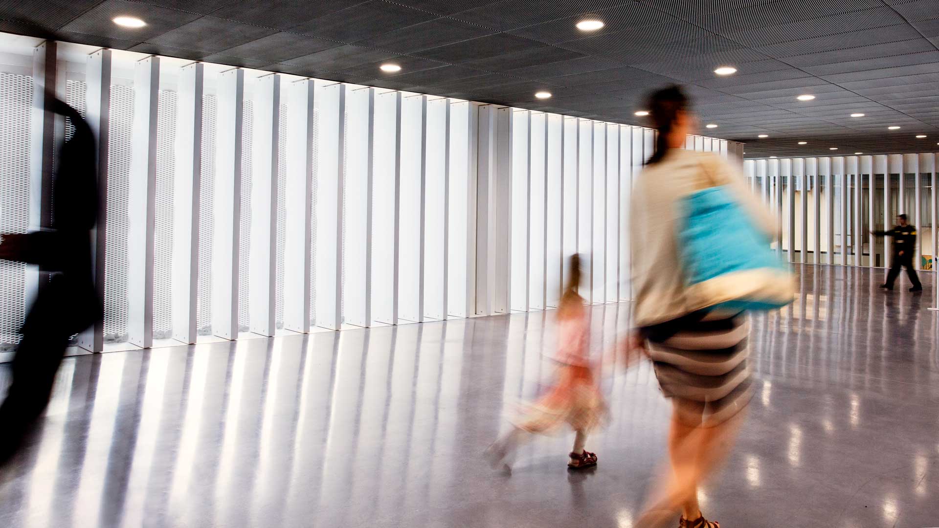 Ein moderner Innenraum mit glatten, weißen vertikalen Lamellen an den Wänden und einer reflektierenden Bodenfläche. Mehrere Personen, darunter ein Kind, bewegen sich unscharf durch den Raum.