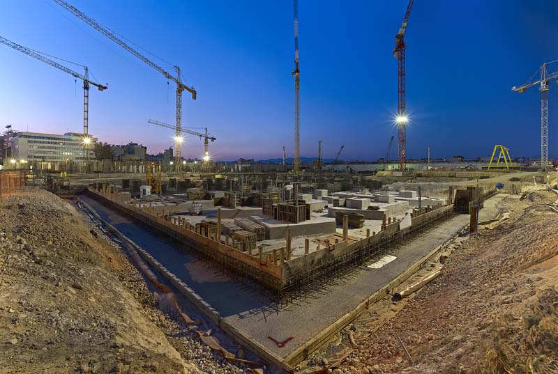 A construction site during twilight with multiple cranes and buildings in the background. The foreground features an excavated area with visible concrete foundations and steel reinforcements, indicating an early stage of construction.