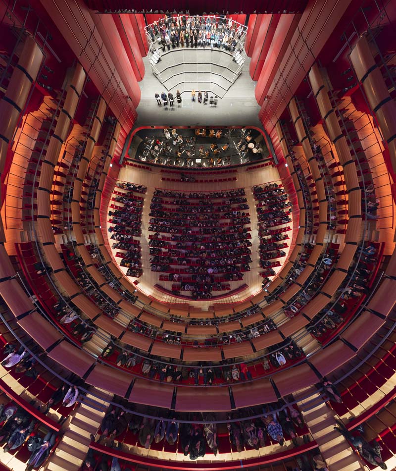 An interior view of a large, circular auditorium with multiple tiers of seating. The seats are arranged in a semi-circular pattern around a central stage area