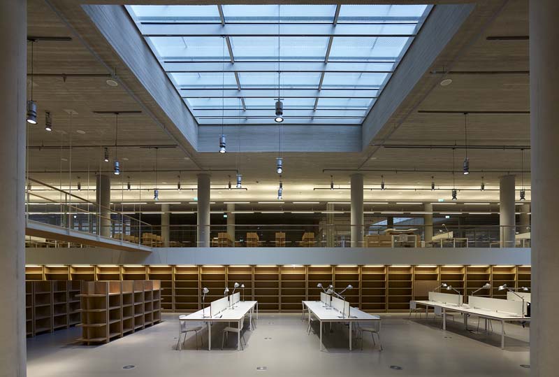 An interior space resembling a library or study area, featuring empty bookshelves, white tables with chairs, and a large skylight providing natural light. The architecture includes exposed beams and ductwork, giving the space an industrial yet modern feel.