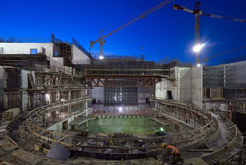 A large construction site during twilight or night, illuminated by artificial lighting. At the center is a circular structure, likely the foundation or lower levels of a building