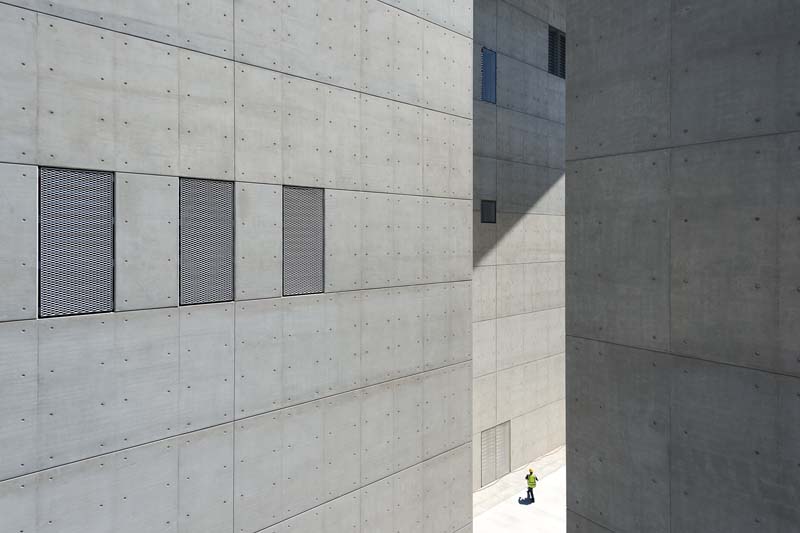 A section of a building with a modern design, featuring a concrete wall with several rectangular recesses and a slightly ajar door on the right side. The play of light and shadow creates an interesting geometric pattern on the surfaces.