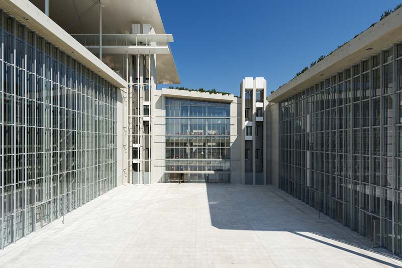 A modern architectural structure featuring a pathway flanked by two buildings with glass facades and geometric designs. The sky is clear, suggesting a bright sunny day.