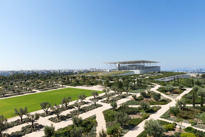 A large, open outdoor space with manicured green lawns and organized rows of trees. In the distance, there is a modern building with a unique, box-like roof structure that appears to be elevated above the ground on pillars or supports.