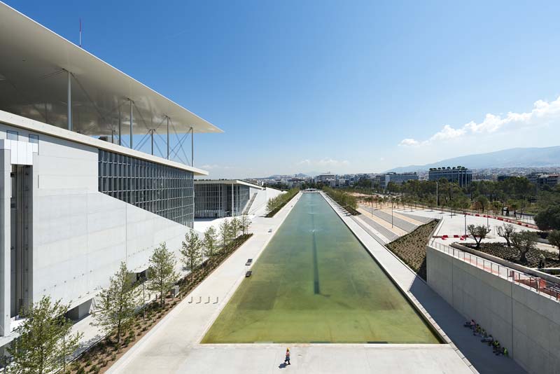 A modern architectural structure featuring a long, narrow water feature extending towards the horizon, flanked by walkways and green spaces. The building has a sleek design with clean lines and expansive glass panels, suggesting it may be a public or commercial space designed for leisure or business activities.