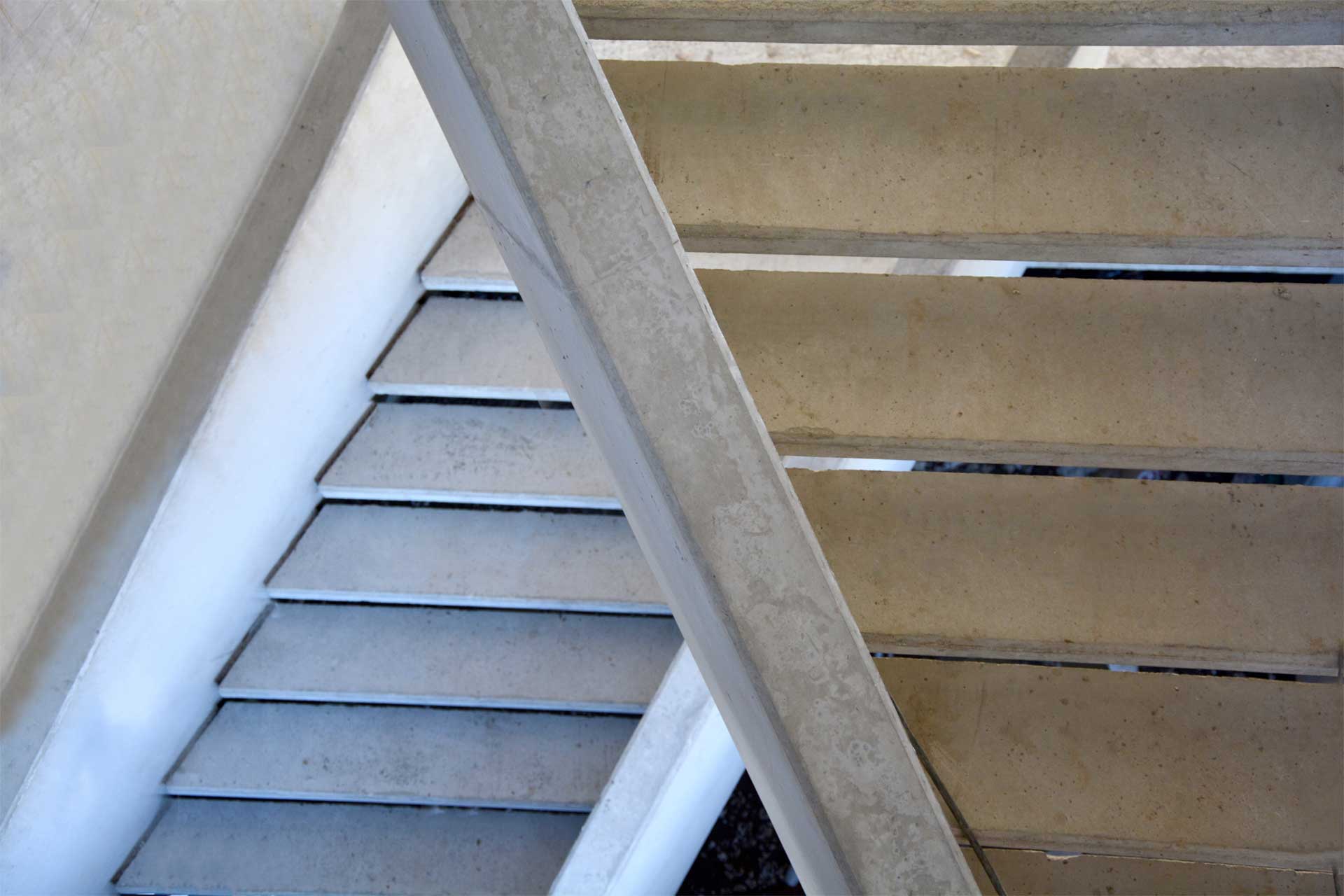 Eine Nahaufnahme eines Treppenabschnitts aus einem schrägen Blickwinkel, die sich auf die Stufen und die Unterseite der Treppe konzentriert. Die Stufen scheinen aus Beton oder einem ähnlichen Material zu bestehen, mit einem Kontrast zwischen dem Licht auf der Oberseite der Stufen und dem Schatten darunter.