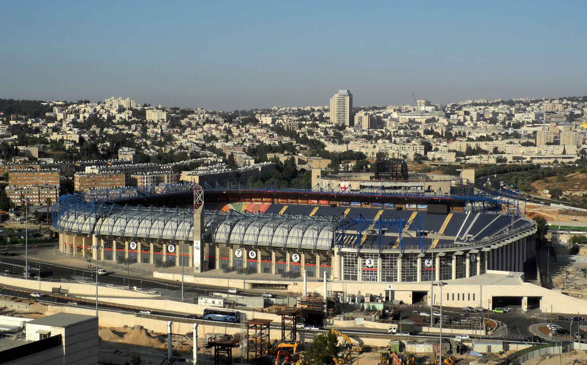 An aerial view of a large stadium with distinctive blue seating and a partial roof structure. Surrounding the stadium is an urban landscape with numerous buildings of varying sizes and designs, roads, and some construction activity in the foreground.