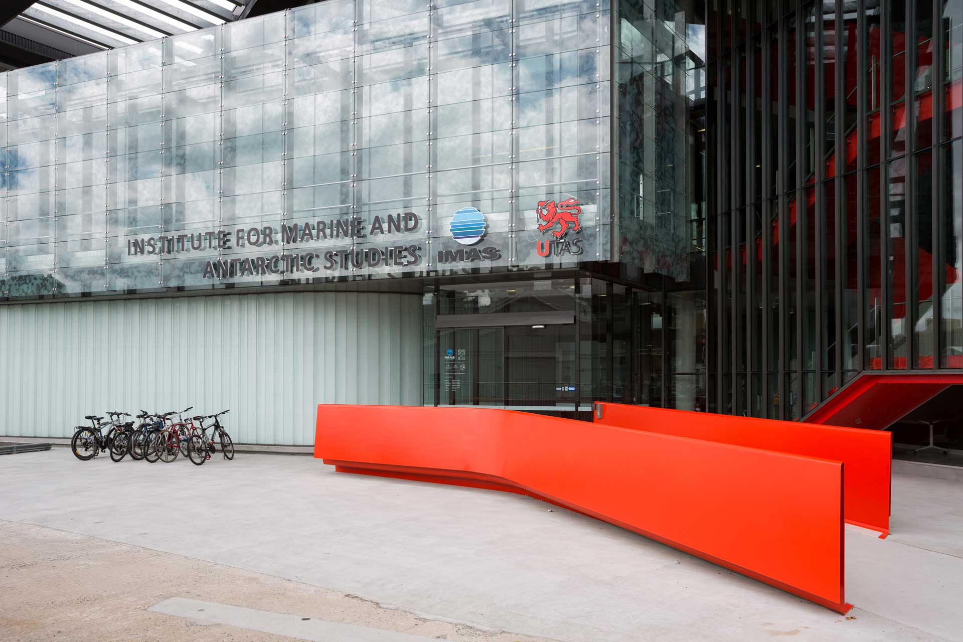 Die Außenansicht eines modernen Gebäudes mit großen Glasfronten und einem Schild mit der Aufschrift “INSTITUTE FOR MARINE AND ANTARCTIC STUDIES” sowie Logos daneben. Vor dem Gebäude befindet sich eine leuchtend orangefarbene, geschwungene Bankstruktur, und links sind Fahrräder in einem Fahrradständer abgestellt.