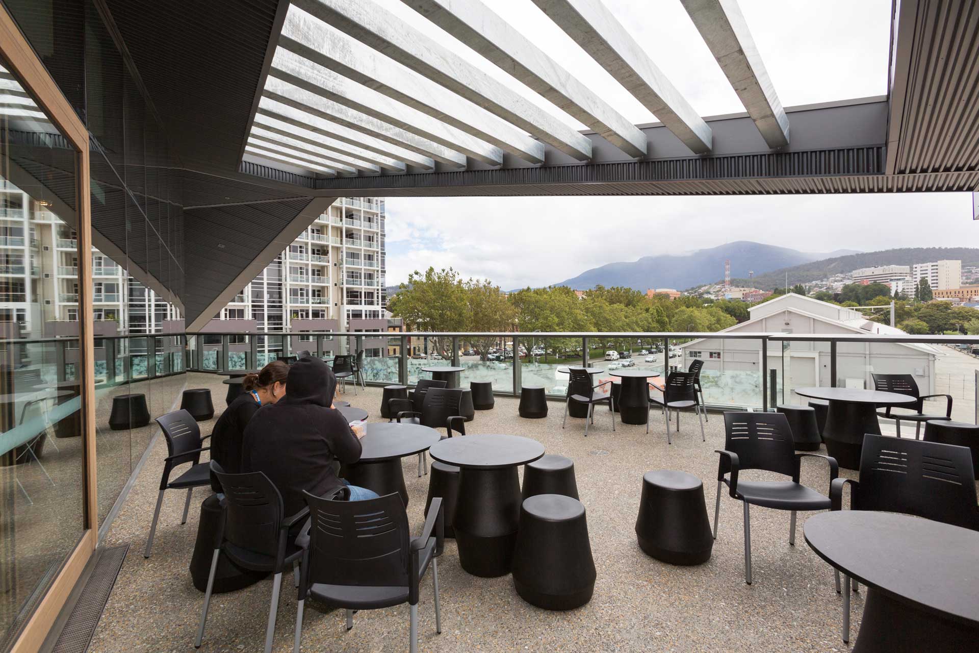 Ein Außenbereich mit Überdachung, ausgestattet mit schwarzen Stühlen und runden Tischen, bietet Blick auf bewaldete Berge und Gebäude. Zwei Personen sitzen an einem Tisch.