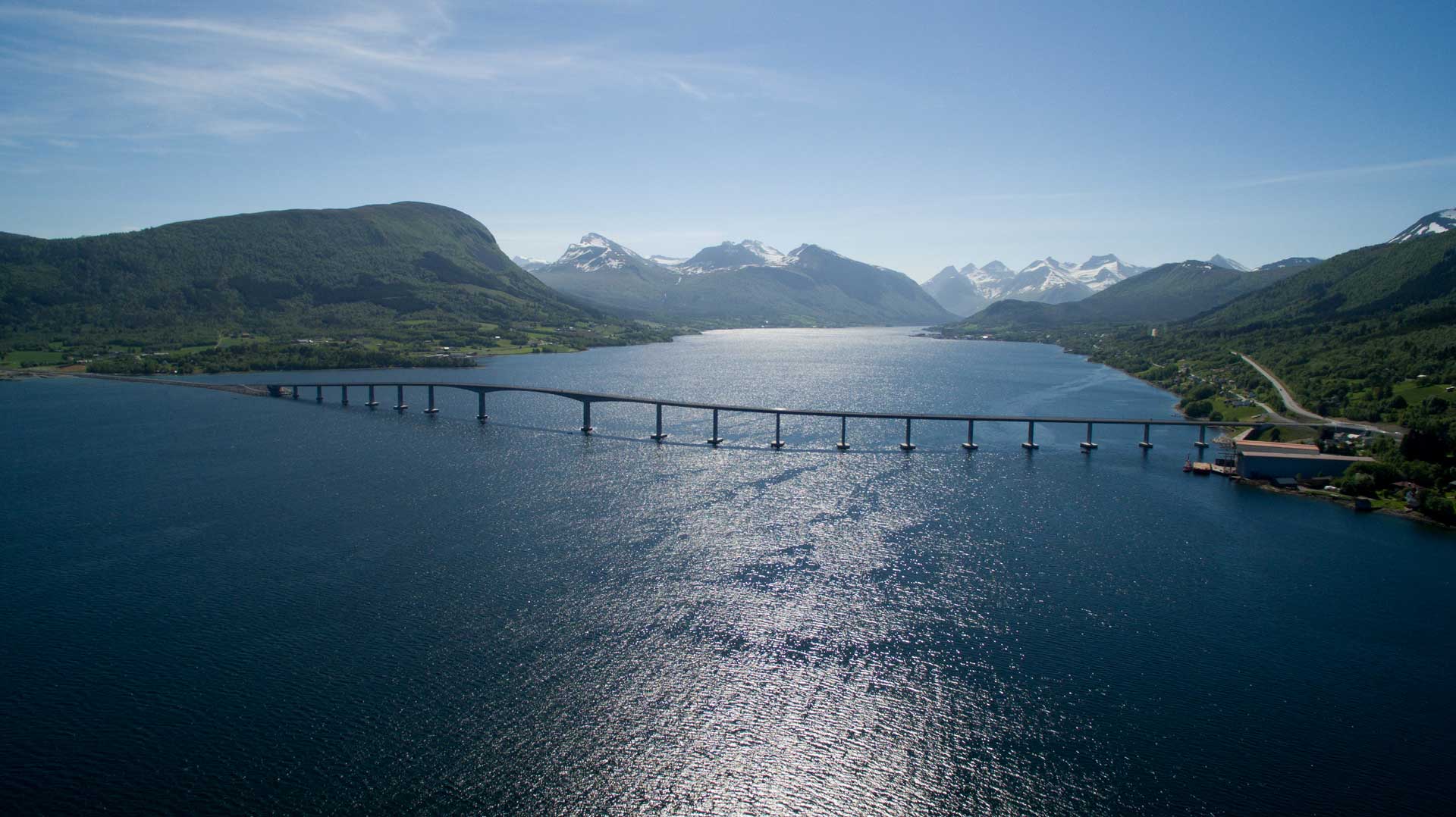 Eine Brücke überspannt eine breite Wasserfläche, umgeben von grünen Ufern und Bergen im Hintergrund. Der Schatten der Brücke zeichnet sich deutlich auf der Wasseroberfläche ab.