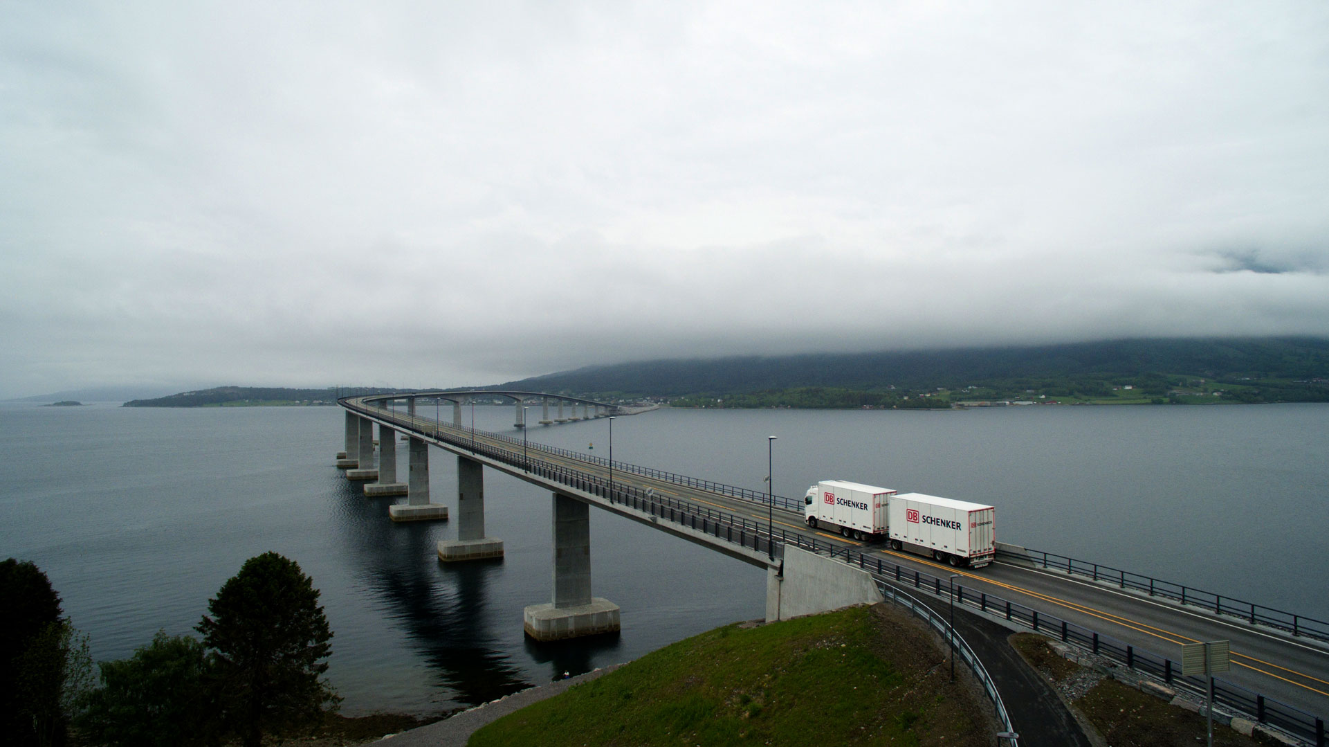 Eine lange Brücke erstreckt sich über ein Gewässer, auf der ein Lastwagen fährt. Die Brücke verbindet zwei Landmassen, und das Wetter ist bewölkt mit tief hängenden Wolken oder Nebel.