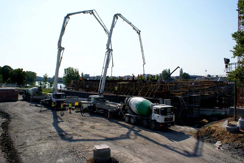 Construction site with a concrete pump truck and its boom extended over a partially constructed structure. A ready-mixed cement truck is also visible, with scaffolding and temporary structures in the background.