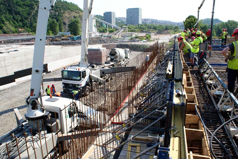 Construction site with several workers wearing safety vests and hard hats. A large crane in the foreground and a ready-mixed concrete truck in the middle ground. Extensive steel reinforcement bars being assembled, with buildings and greenery on a hillside in the background under a clear sky.