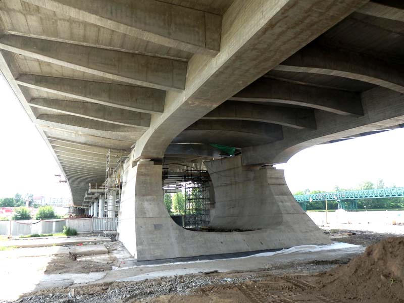 Underside of a large concrete bridge with multiple support columns and visible scaffolding on one side, indicating construction work. Ground beneath is a mix of dirt and gravel, with a green fence in the background.