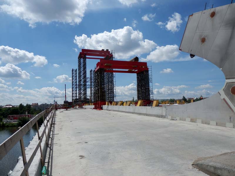 Construction site for a bridge with large red gantry cranes and multiple black scaffolding structures. Partly cloudy sky with buildings visible in the distance.