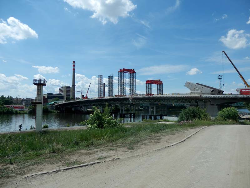 Bridge under construction over a body of water with multiple cranes and scaffolding structures. Partly cloudy sky, dirt path in the foreground leading towards the bridge. Industrial building with a tall chimney and two spherical structures on towers on the left.