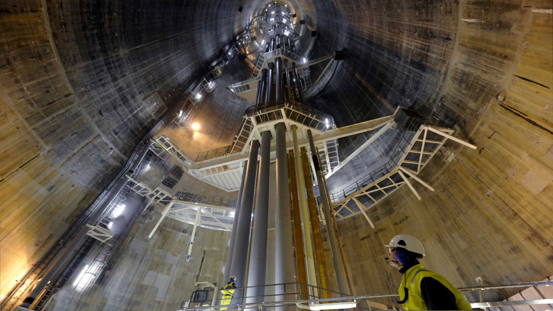 Das Innere der Bohrinsel Troll A, einer großen zylindrischen Struktur mit mehreren Metallträgern und Gerüsten. Ein Arbeiter mit Schutzhelm und reflektierender Weste ist im Vordergrund zu sehen, was auf eine industrielle oder bauliche Umgebung hinweist.