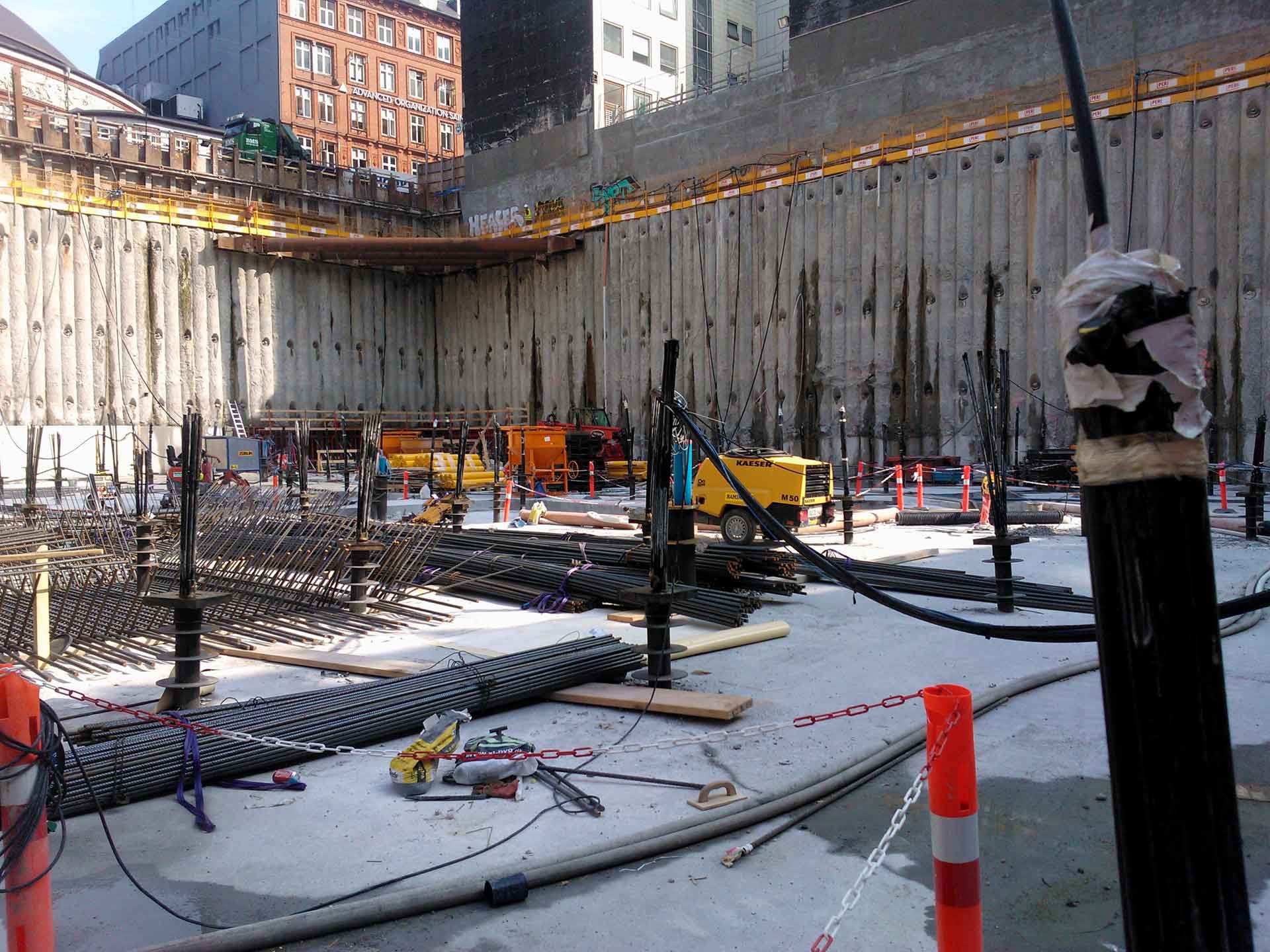 Eine Baustelle mit einer großen Baugrube. Es gibt verstärkte Stahlstangen auf dem Boden, schwere Maschinen wie einen Bagger und einen Kran sowie verschiedene Baumaterialien, die verstreut herumliegen.