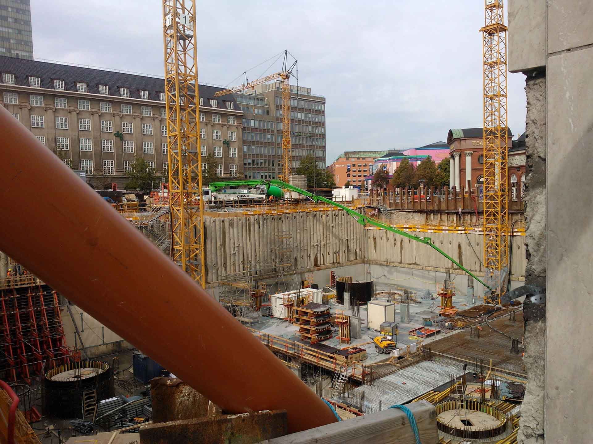 Eine Baustelle mit mehreren Kränen, einer tiefen Baugrube mit Stützwänden und verschiedenen Baumaterialien, die auf dem Gelände verstreut sind. Im Hintergrund sind Gebäude zu sehen, was darauf hinweist, dass diese Baustelle in einem städtischen Gebiet liegt.