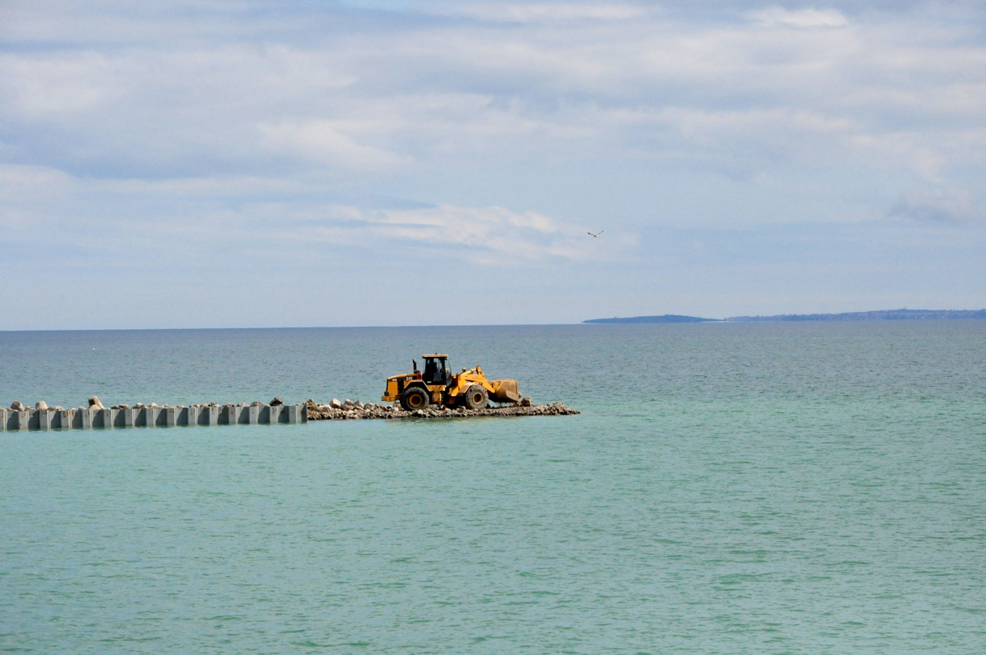 A yellow bulldozer is working on a small, man-made island in a large body of water. There are rocks and a breakwater around the island.