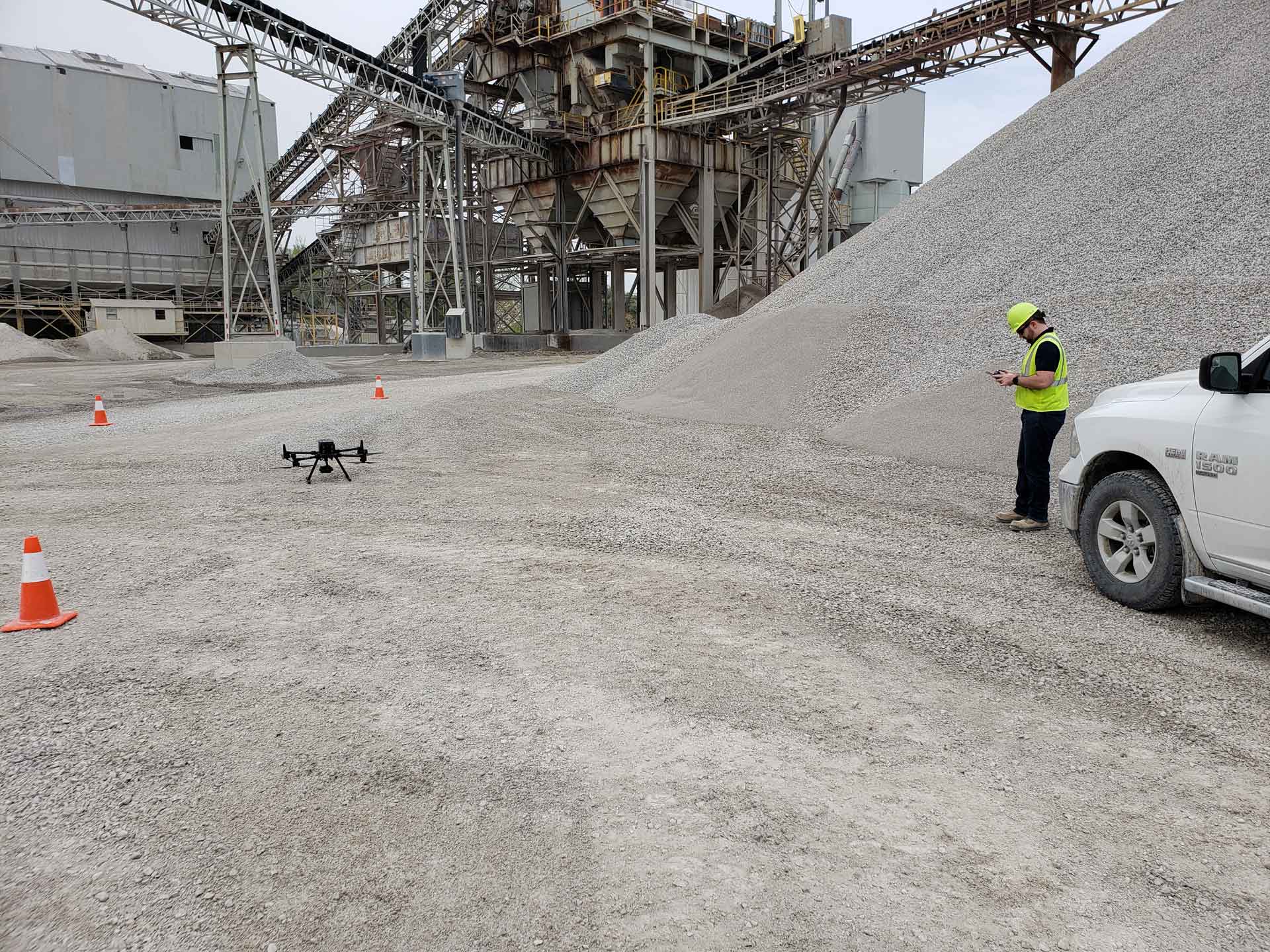 A drone stands on a gravel area in a quarry. In the background are large piles of gravel, industrial plants and conveyor belts. On the right is a person in protective clothing controlling the drone.