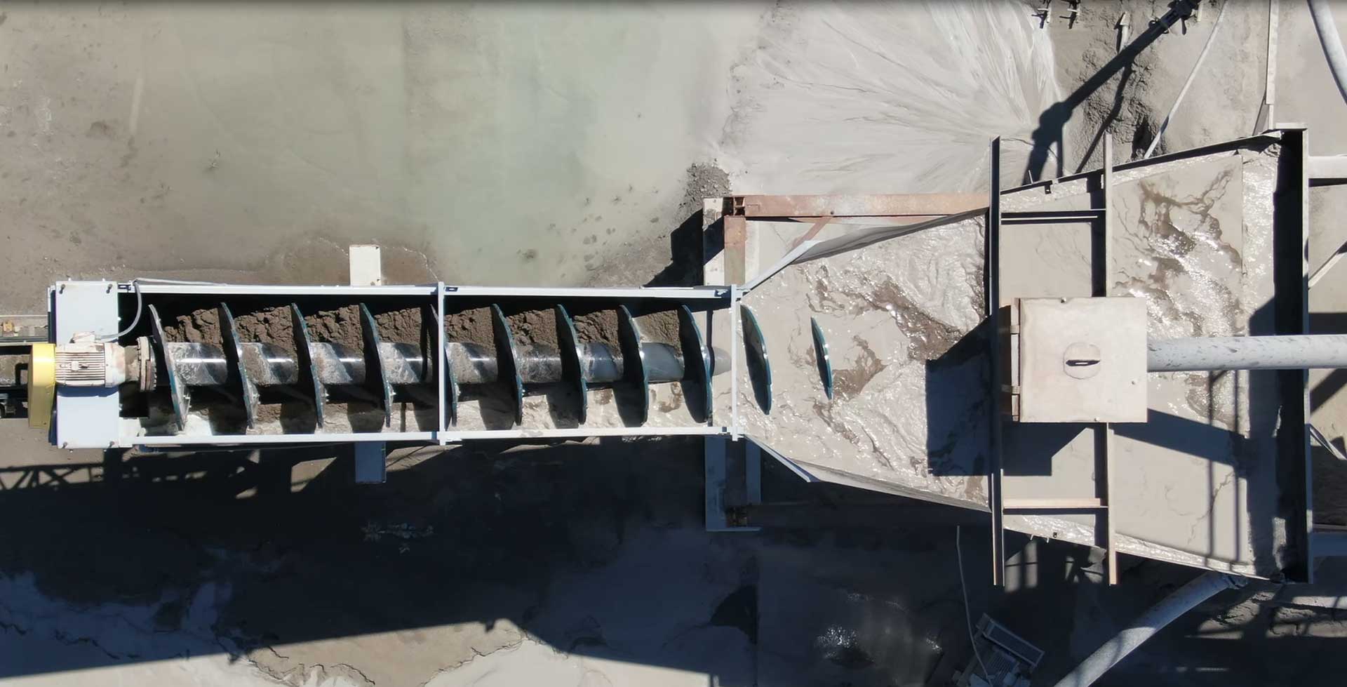 An aerial top-down view of a large industrial spiral conveyor belt. The belt is filled with a dark, muddy substance and is discharging it onto a concrete surface below.