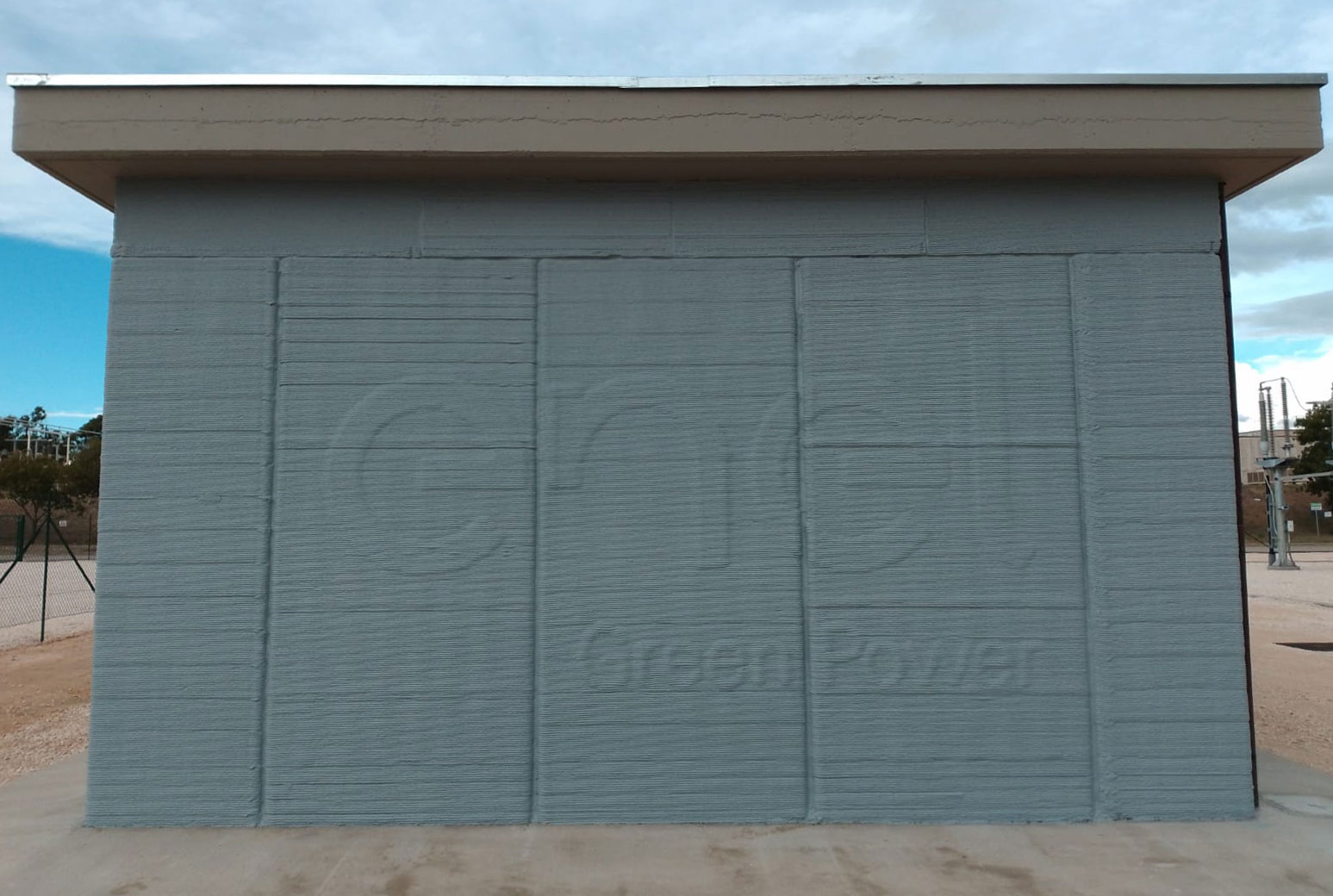 A 3D-printed side wall of a building, showcasing the unique layered texture characteristic of 3D printing technology. The wall appears robust and is set against a backdrop of a cloudy sky. The imprint "Enel Green Energy" is visible.