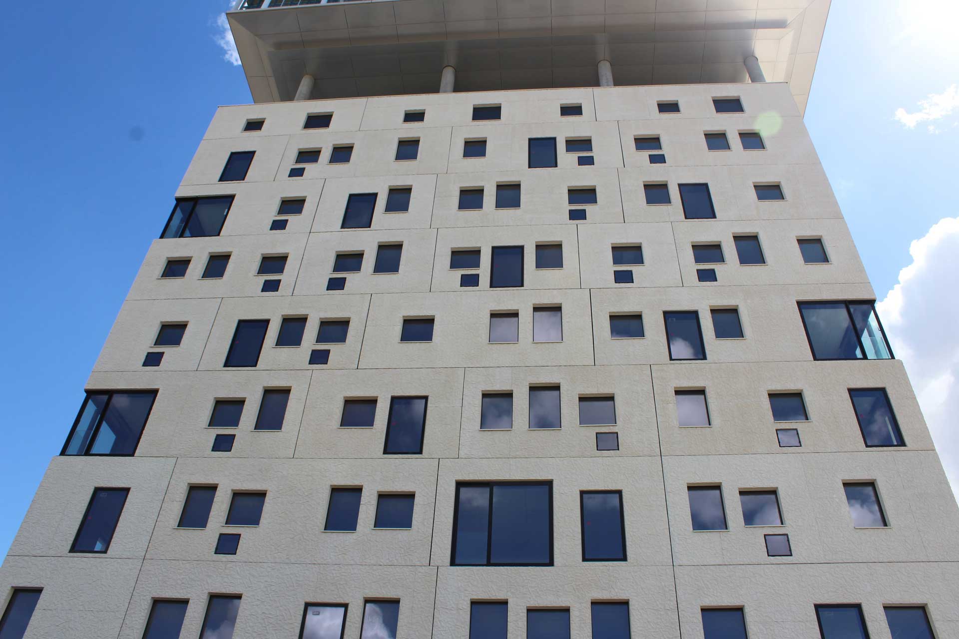 A building that consists of two cubes that are built above each other, the upper cube positioned slightly offset to the side. The lower building’s facade is smooth, with embedded windows of different shapes and sizes while the upper building has a regular, glazed facade